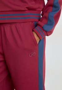 Bordeaux tracksuit med blå detaljer, som har striber på ærmerne og taljen. Inkluderer en lomme og et logo på siden af bukserne.
