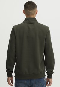 Sudadera verde oliva con un alto cuello, mangas largas y puños de canalé. Tejido suave con costuras mínimas y sin logotipos visibles.
