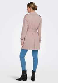 Trench-coat rose clair avec une taille ceinturée, des épaulettes et des poignets boutonnés, associé à un jean skinny bleu et des bottines noires.
