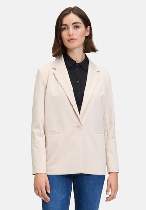 Light beige blazer with a single button closure, notch lapels, and two front pockets. Worn over a black collared shirt and blue jeans.