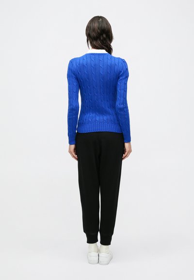 Kék, kötött mintás pulóver bordázott szegéllyel, fekete, szűkített szárú melegítőnadrággal és fehér tornacipővel, hátulnézetben.