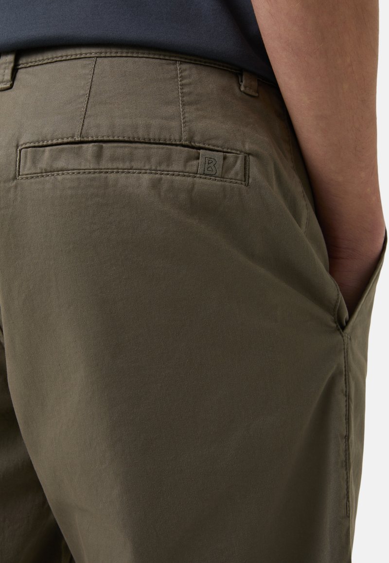 Olivgrüne Hose aus glattem Stoff. Sie verfügt über eine Gesäßtasche mit einem gestickten Emblem und hat einen klassischen Schnitt mit einem dezenten Glanz.