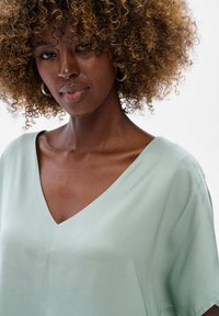 Miętowo-zielona bluzka z dekoltem w serek i krótkimi rękawami, o gładkiej teksturze i subtelnych detalach ściegów. Posiada luźny krój i płynny spadek.
