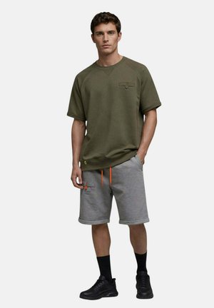 Homme portant un sweat-shirt à manches courtes vert olive, un short gris arrivant aux genoux avec des cordons orange, des chaussettes noires et des baskets noires, debout la main dans la poche.