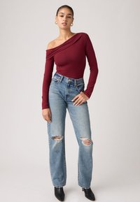 Γυναίκα με μπορντό τοπ με ένα ώμο και μακριά μανίκια, σκισμένο μπλε τζιν, μαύρα μπότες με τακούνι και χρυσούς κρίκους ως σκουλαρίκια, μπροστά σε απλό φόντο.