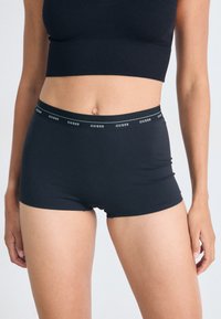 Shorts stretch noirs en tissu doux avec une taille large arborant le logo "GUESS" en blanc. Texture lisse et design ajusté.