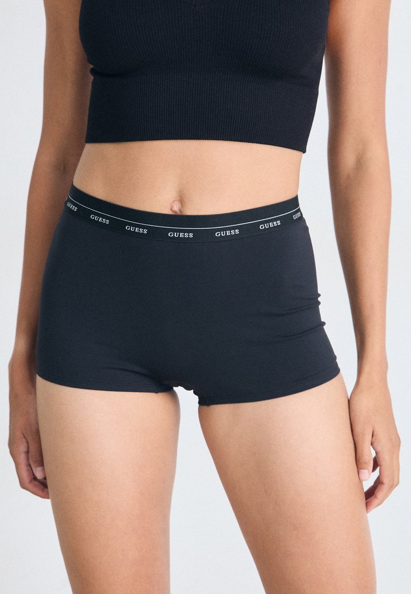 Shorts stretch noirs en tissu doux avec une taille large arborant le logo "GUESS" en blanc. Texture lisse et design ajusté.