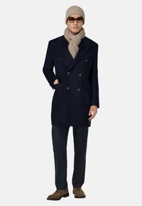 Abrigo navy de doble botonadura con grandes botones, combinado con una bufanda y un gorro de punto beige; unos pantalones texturizados y unos zapatos de cordones marrones completan el look.
