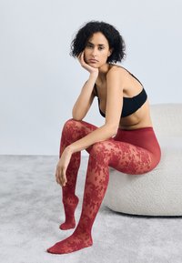 Rote florale Spitzenstrumpfhosen aus transparentem Material mit hoher Taille. Das Model posiert sitzend auf einer strukturierten, neutralen Oberfläche.