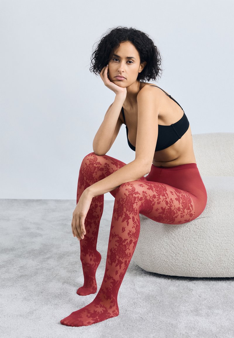 Rote florale Spitzenstrumpfhosen aus transparentem Material mit hoher Taille. Das Model posiert sitzend auf einer strukturierten, neutralen Oberfläche.