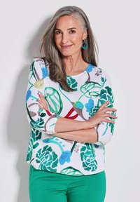 Driekwart mouw shirt in het wit met een kleurrijk bloemenpatroon in groen, blauw en rood. Draagt met groene broek en heeft een ronde hals.