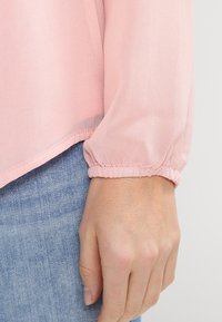 Blouse rose, légère et ample, avec des manches bouffantes et des poignets élastiques, assortie à un denim bleu clair.