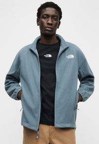 Ζακέτα από φλις σε απαλό γαλάζιο χρώμα με φερμουάρ στο μπροστινό μέρος, ψηλό γιακά και λεπτομέρειες με λογότυπο. Φοράται πάνω από μαύρο μπλουζάκι με παρόμοιο λογότυπο και μπεζ παντελόνι.