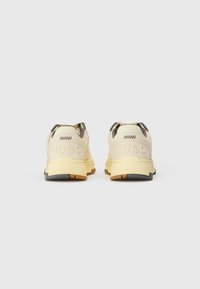 Zapatillas deportivas de color beige claro con parte superior texturizada, acentuadas con gris y marrón oscuro. Marca "HUGO" grabada en el talón. Suela de goma.