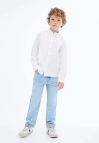 Chemise blanche à manches longues, jean bleu clair et baskets beiges avec des accents. L'enfant se tient avec une main dans la poche contre un fond uni.