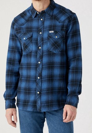 Homme portant une chemise à carreaux bleue et noire à manches longues avec deux poches poitrine et un jean bleu sur un fond uni.