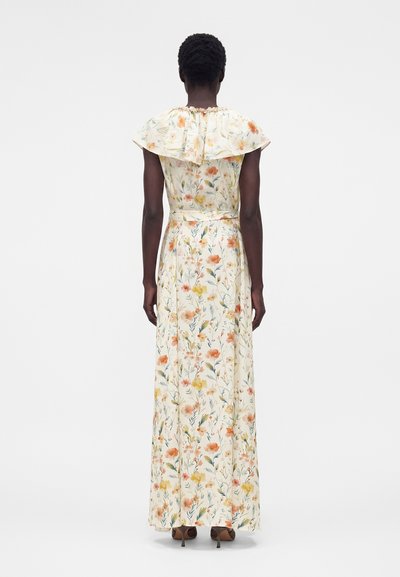 Γυναίκα που στέκεται με την πλάτη προς το φακό, φορώντας μακρύ κρεμ φόρεμα με λουλουδένιο μοτίβο, με γιακά με βολάν και καφέ ψηλοτάκουνα παπούτσια σε απλό φόντο.