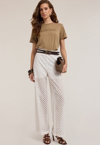 Camiseta marrón con el texto "HAZ HOY", combinada con pantalones anchos blancos perforados y un cinturón oscuro. Los accesorios incluyen un bolso de mimbre y pulseras.