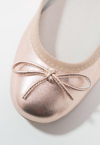 Bailarina en rosa dorado metálico, con puntera redondeada y un detalle de lazo decorativo en la parte frontal. Parte superior suave y flexible con bordes elastizados.