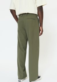 Olívazöld, pamutkeverékből készült nadrág; egyenes szárú kialakítás hátsó zsebekkel és övbújtatókkal, fekete-fehér sportcipővel párosítva.