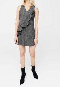 Graues ärmelloses Kleid mit V-Ausschnitt und diagonalem Rüschendetail auf der linken Seite; kombiniert mit schwarzen Stiefeletten.