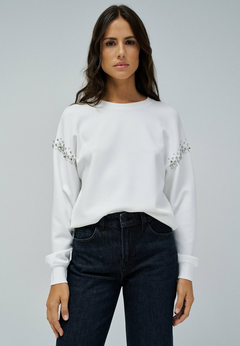 Sweatshirt blanc à manches longues avec des embellissements sur les épaules, associé à un jean en denim foncé. Texture lisse, coupe décontractée.