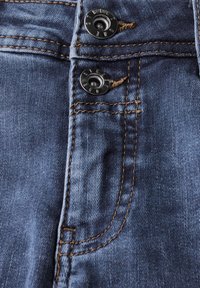 Jeans en denim bleu foncé avec deux boutons en métal noir et des coutures dorées contrastantes, avec une poche avant visible sur la gauche.