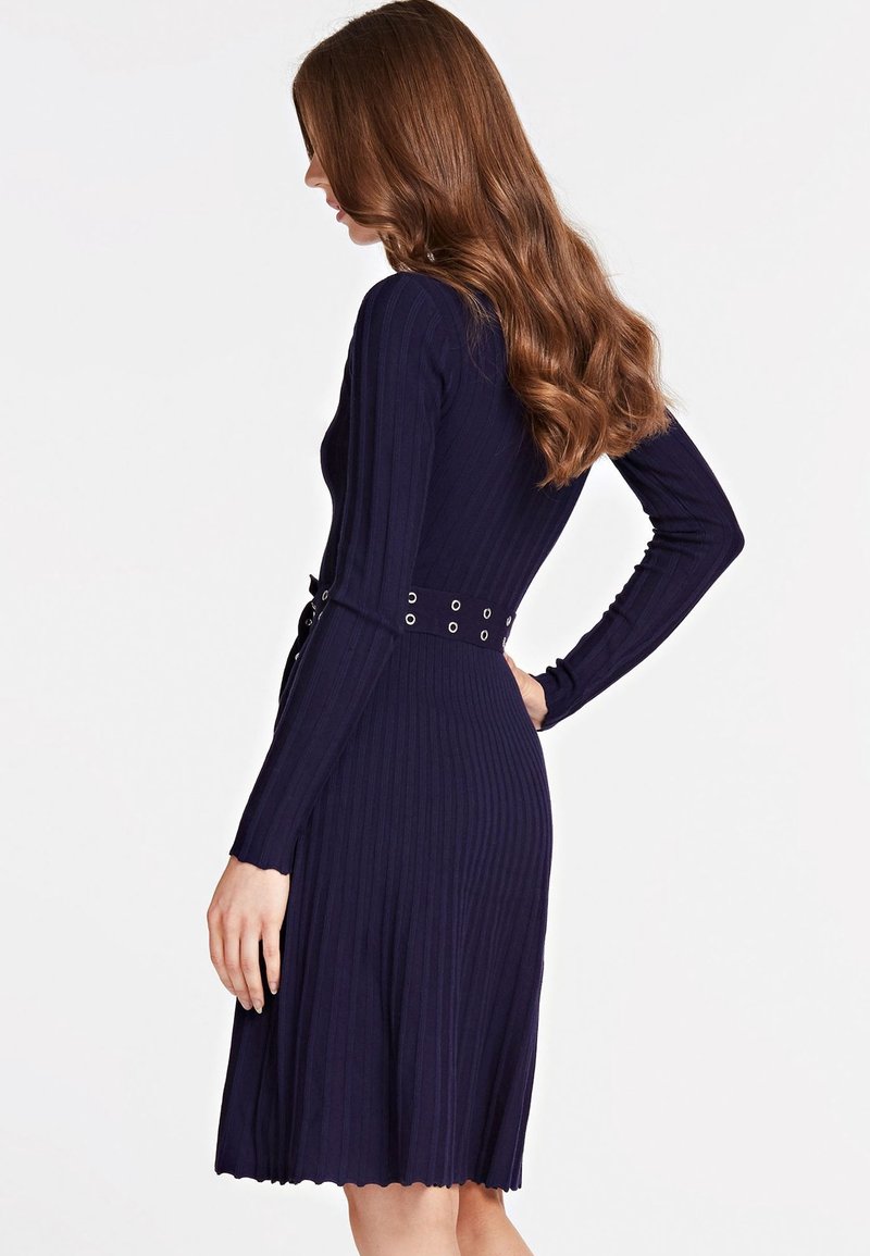 Femme aux longs cheveux roux portant une robe en maille côtelée bleu marine à manches longues avec une ceinture à la taille, vue de profil et de dos.
