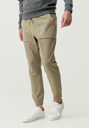 Person trägt beige Jogginghosen mit Fronttaschen und weißen Schnürsneakern, steht vor einem einfarbigen Hintergrund.