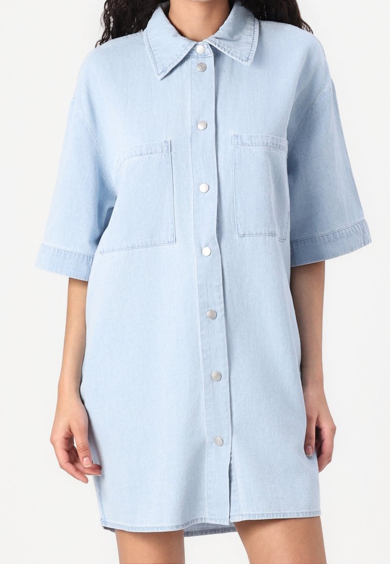 Robe chemise en denim bleu clair avec manches courtes, boutonnage à l'avant, deux poches poitrine et un col à revers. Texture lisse et coupe décontractée.
