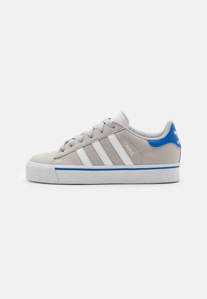 Beige Adidas Campus sneaker med hvide striber, blå hælflap og kant, hvid sål og snørrebånd på hvid baggrund.