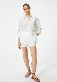 Camisa blanca de botones con sutiles patrones de hojas, mangas enrolladas y un ajuste relajado, combinada con pantalones cortos blancos y sandalias marrones de tiras.