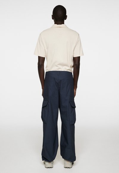 Beige Poloshirt mit kurzen Ärmeln, marineblaue Cargohose mit großen Seitentaschen und grauen Sneakers. Glattes Material und lockere Passform.
