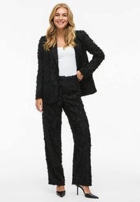 Black textured suit with a fitted blazer and straight-leg pants, made from a fabric with a fringe detail, paired with a white top and heels.