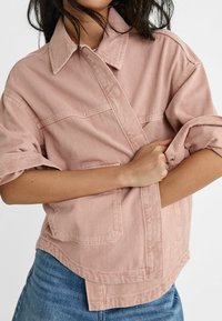 Camicia di jeans rosa chiaro con colletto a punta, maniche arrotolate, due tasche sul petto e taglio dritto, caratterizzata da un tessuto intrecciato dalla texture evidente.