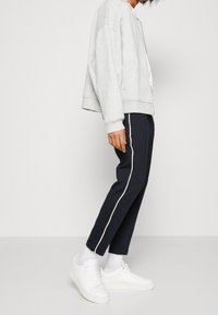 Sweatshirt zippé gris avec une coupe relax, associé à un pantalon bleu marine avec des rayures blanches sur les côtés et des baskets blanches à lacets.