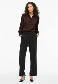 Camicia lunga in satin marrone con colletto, abbinata a pantaloni neri a vita alta. Il modello indossa tacchi neri a punta.