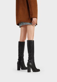 Stivali neri fino al ginocchio in pelle liscia con tacco spesso, dotati di cerniera laterale e sottili accenti con fibbia.