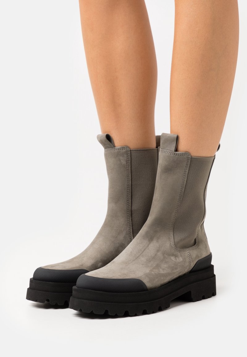 Bottes mi-mollet en daim gris clair avec semelles noires robustes portées sur jambes nues, avec languettes de tirage et panneaux latéraux élastiques.