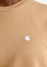 Hnědý bavlněný tričko s žebrovaným výstřihem do kulata. Obsahuje malé bílé logo na hrudníku. Měkká textura, jednoduchý design, žádné vzory.