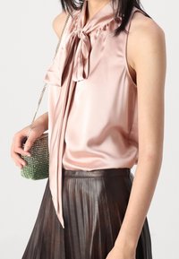 Blusa di seta rosa con un dettaglio al colletto a fiocco, abbinata a una gonna plissettata marrone. Tiene in mano una piccola borsa in strass con texture ombré verde.