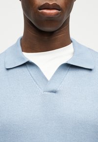 Κοντινό πλάνο ενός ατόμου που φοράει ένα ανοιχτό μπλε πλεκτό πουλόβερ πόλο πάνω από ένα λευκό μπλούζα με στρογγυλή λαιμόκοψη, παρουσιάζοντας το λαιμό και τους ώμους.