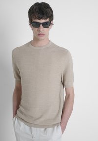 Camiseta de punto beige de manga corta con un sutil patrón texturizado. Presenta un cuello redondo y un dobladillo acanalado, combinada con gafas de sol rectangulares negras.