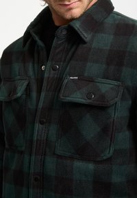 Flannel shirt in dark green and black plaid, featuring two front pockets, snap closures, and a soft texture. Brand logo on pocket.