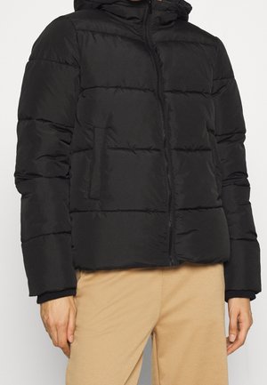 Personne portant une doudoune noire avec fermeture éclair à l'avant et poches, associée à un pantalon beige sur un fond clair uni.
