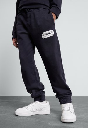 Person iført sorte joggerbukser med et hvidt firkantet logo på låret og hvide New Balance-sneakers på gråt gulv.