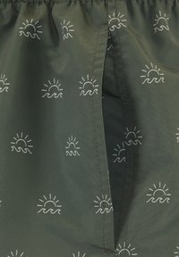 Zelená tkanina s vzorom slnka a vĺn, obsahujúca otvor na vrecko a elastický pás s volánmi. Hladká textúra, ľahký materiál.