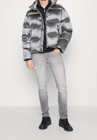 Mand iført en skinnende sølv pufferjakke, sort skjorte, lysegrå slidte skinny jeans og sorte sneakers, som står op ad en hvid baggrund.