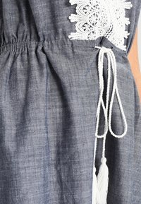 Denimově modré šaty s bílými krajkovými detaily na rameni, staženým pasem se zavazováním z bílého provázku a střapcovými ozdobami.