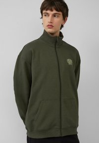 Olivgrüner Reißverschluss-Pullover aus weichem Material. Mit einem hohen Kragen, Taschen vorne und einem kleinen Logo auf der Brust.